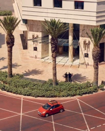 High-angle exterior shot of a hotel entrance with palm trees and a red car parked on a reddish paved roundabout.
