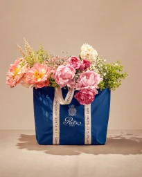 Blue tote bag overflowing with pink and white peonies and wildflowers on a neutral beige background