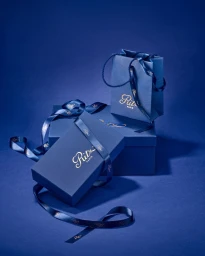 Blue Ritz Paris gift boxes and a paper shopping bag tied with branded satin ribbon on a matching blue studio background