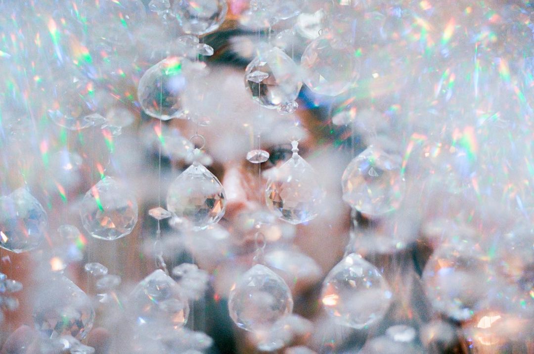 Close-up of a person's face obscured by clear crystal pendants, refracting light and creating a soft, ethereal effect.