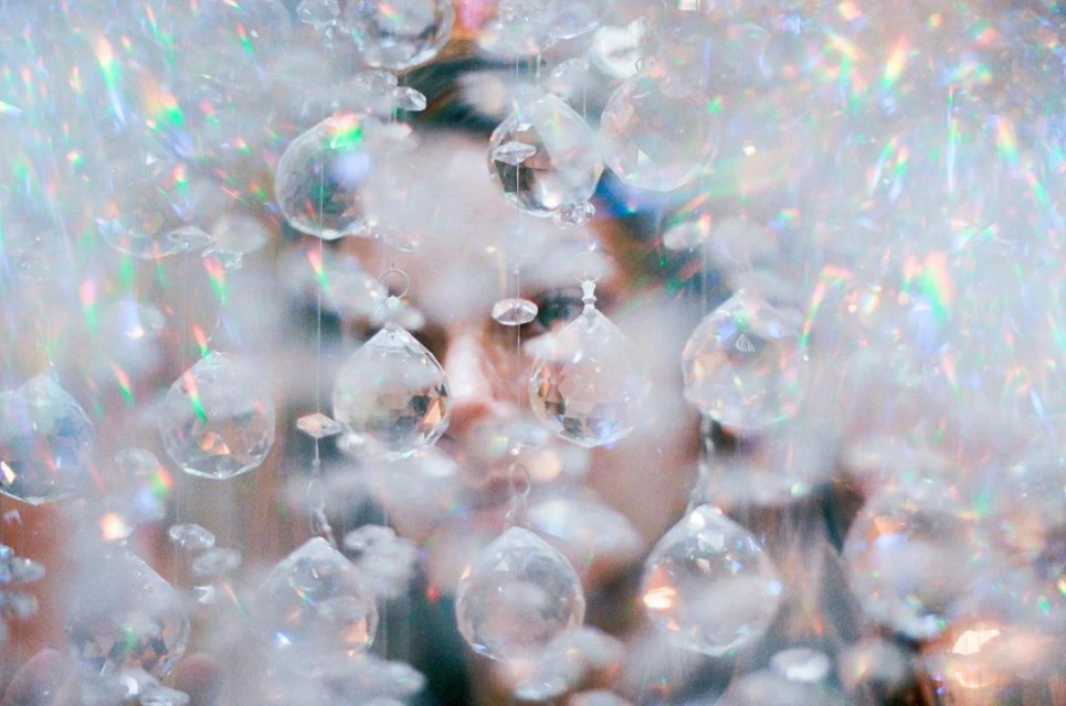 Close-up of a person's face obscured by clear crystal pendants, refracting light and creating a soft, ethereal effect.