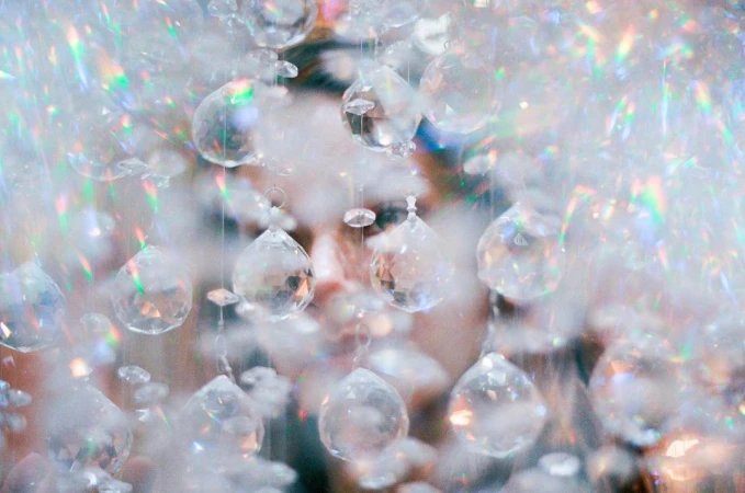 Close-up of a person's face obscured by clear crystal pendants, refracting light and creating a soft, ethereal effect.