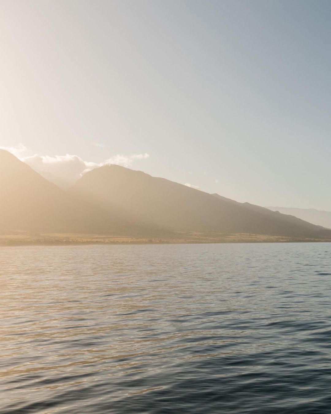 Sunlit mountain range across calm ocean water with warm golden sunlight filtering through clouds.