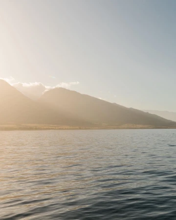Sunlit mountain range across calm ocean water with warm golden sunlight filtering through clouds.