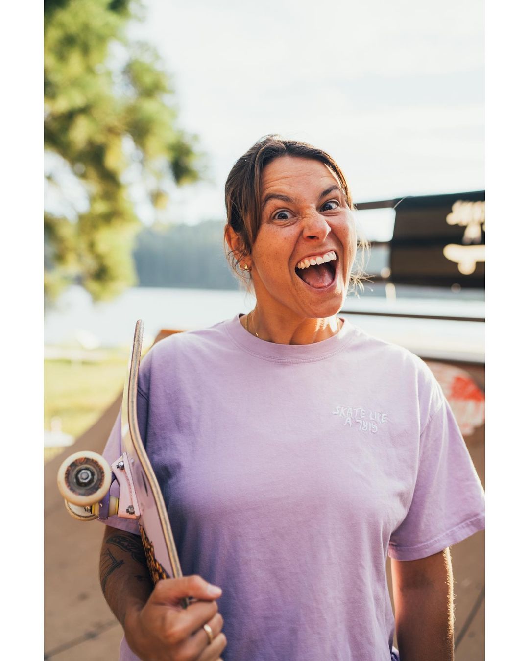 Woman in lilac shirt with skateboard grins at camera outdoors. Trees and a lake are visible in background on bright day.