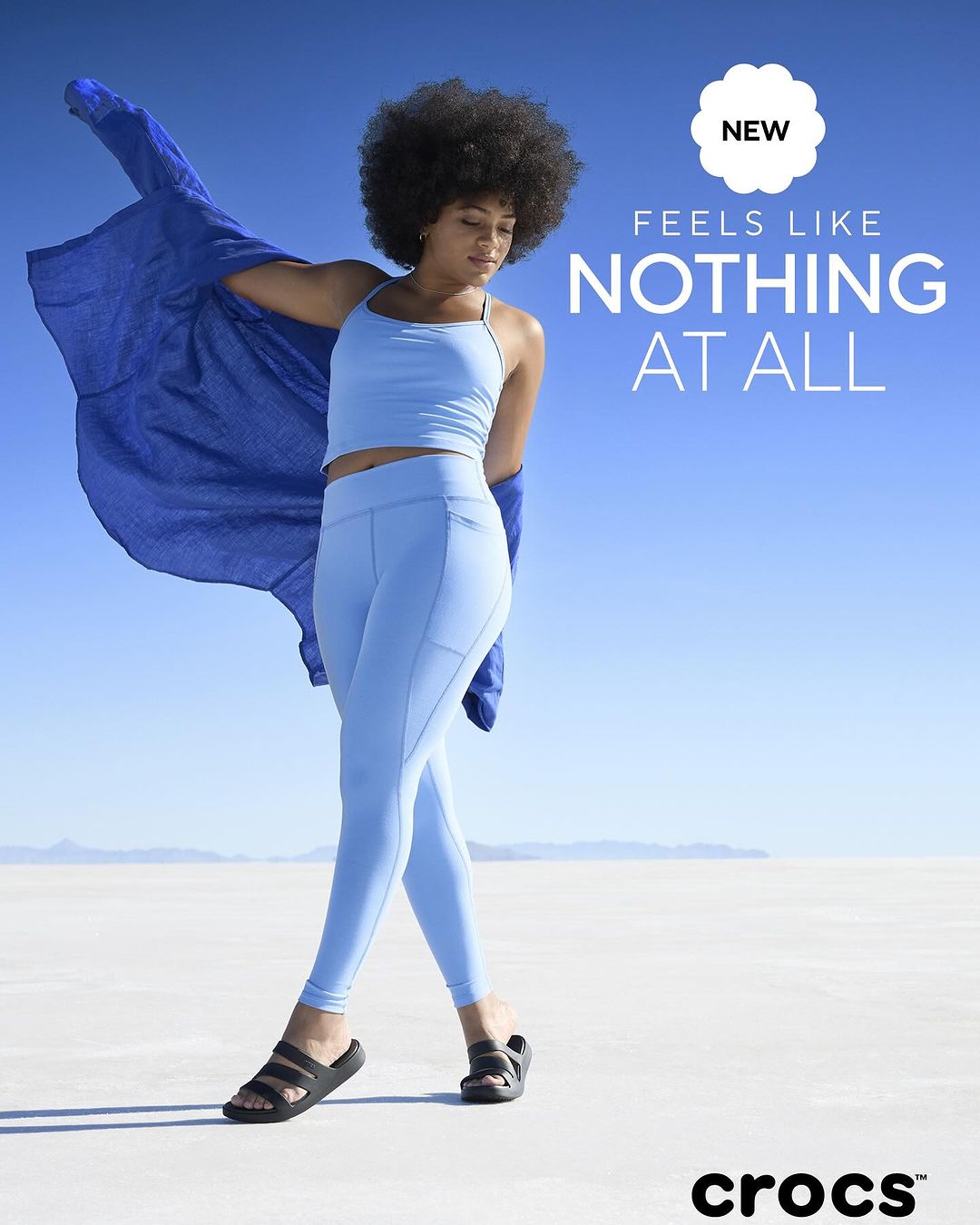 An athletic woman with afro-style hair poses outdoors in light blue workout clothes and black sandals against a white salt flat.