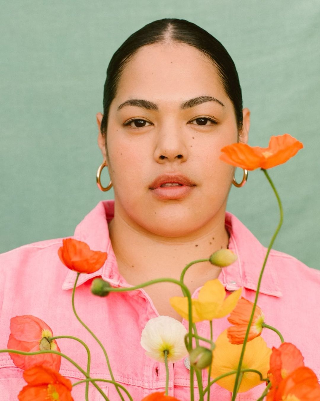 Portrait of an adult female surrounded by a vibrant arrangement of orange, yellow, and white poppies against a muted green backdrop.