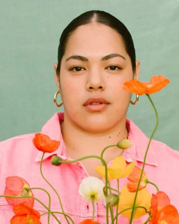 Portrait of an adult female surrounded by a vibrant arrangement of orange, yellow, and white poppies against a muted green backdrop.