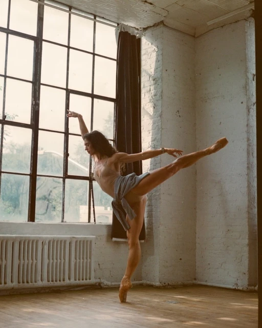 A ballerina in pointe shoes poses in a dance studio with a large window, showcasing her flexibility and form with natural light.