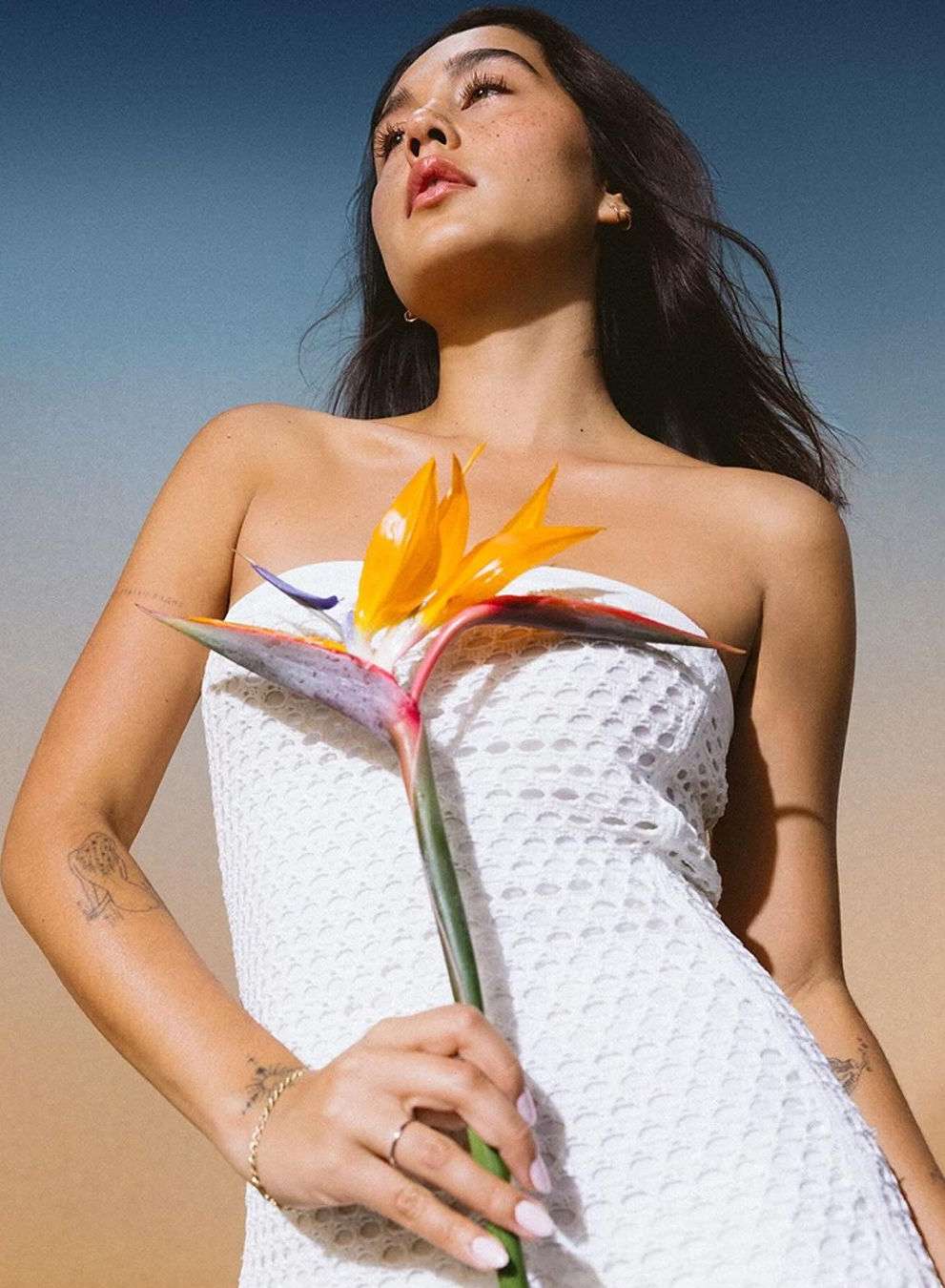 Young woman poses in a white dress outdoors, holding a bird of paradise flower against a sky with tan and blue gradient.