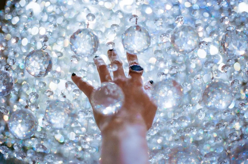 A human hand with a ring extends upward among a cascading waterfall of clear, dangling crystal spheres against a bokeh background.