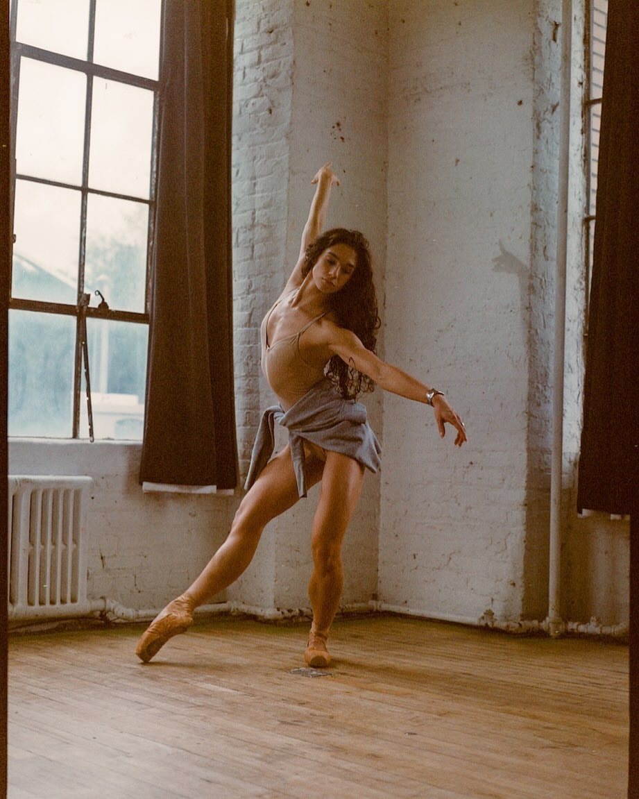 A female ballet dancer in a beige leotard and blue skirt performs in a dance studio with a wooden floor and large windows.