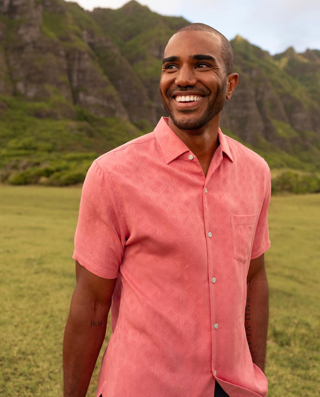Smiling adult man in a salmon short sleeve shirt stands outdoors against a mountain backdrop with green meadow in the foreground.