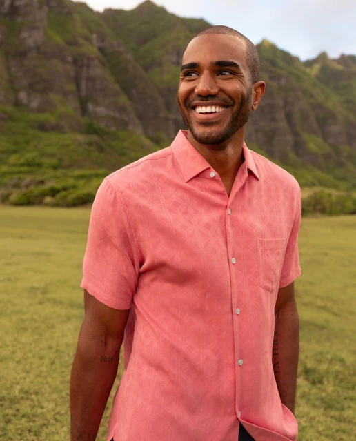 Smiling adult man in a salmon short sleeve shirt stands outdoors against a mountain backdrop with green meadow in the foreground.