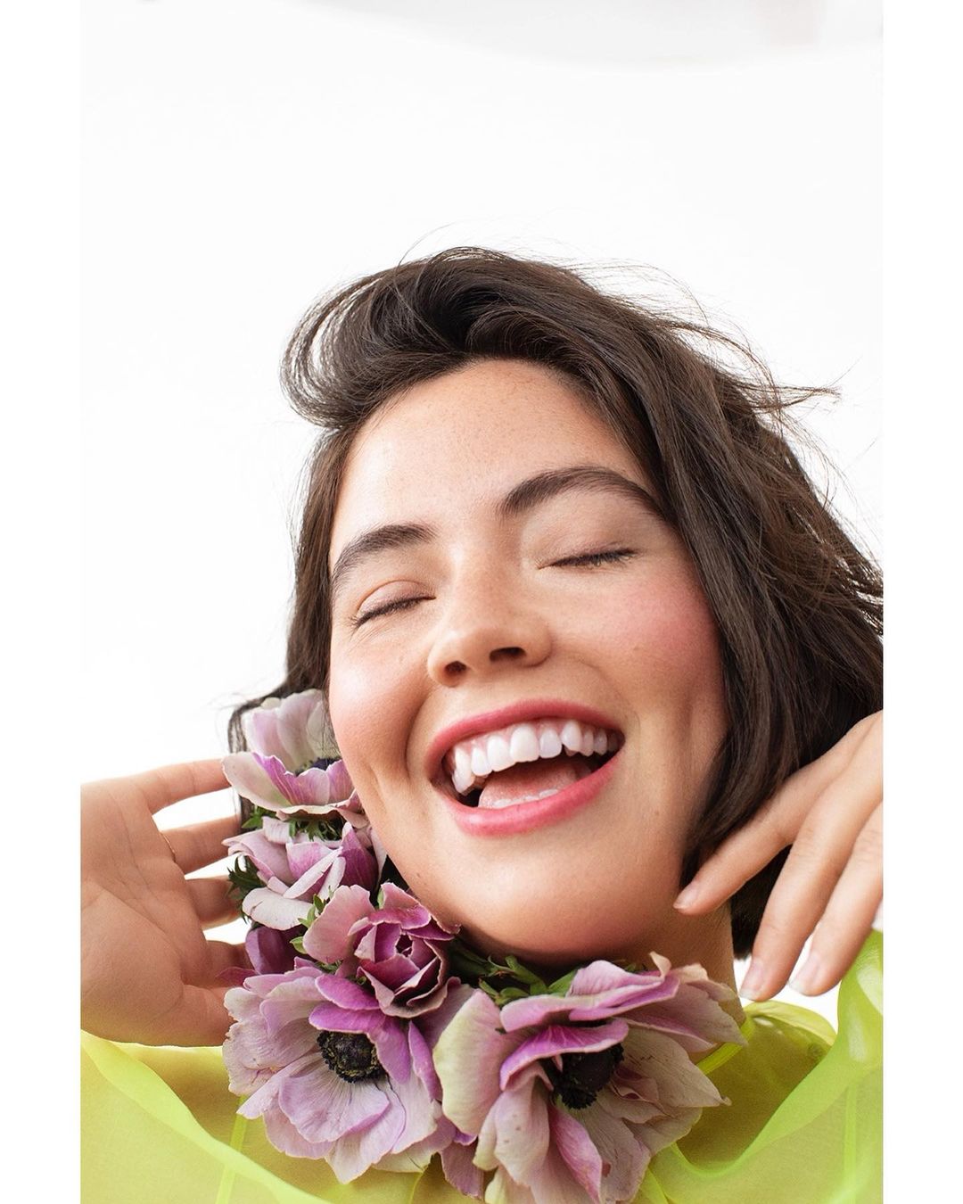 Smiling woman with closed eyes, adorned with pink and purple anemone flowers around her neck, wearing a sheer yellow top.