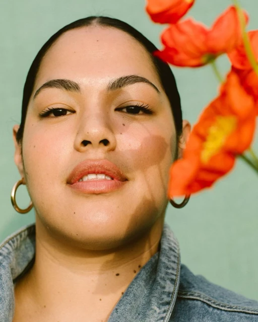 Portrait of a woman with gold hoop earrings and a denim jacket, framed by red and yellow flowers against a light green backdrop.