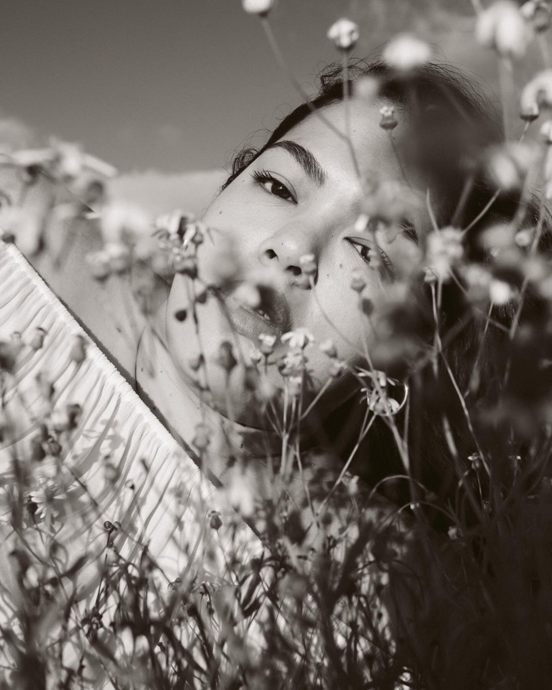 Monochrome portrait of an adult female in a field of flowers, gazing at the camera, with a clear sky in the background.
