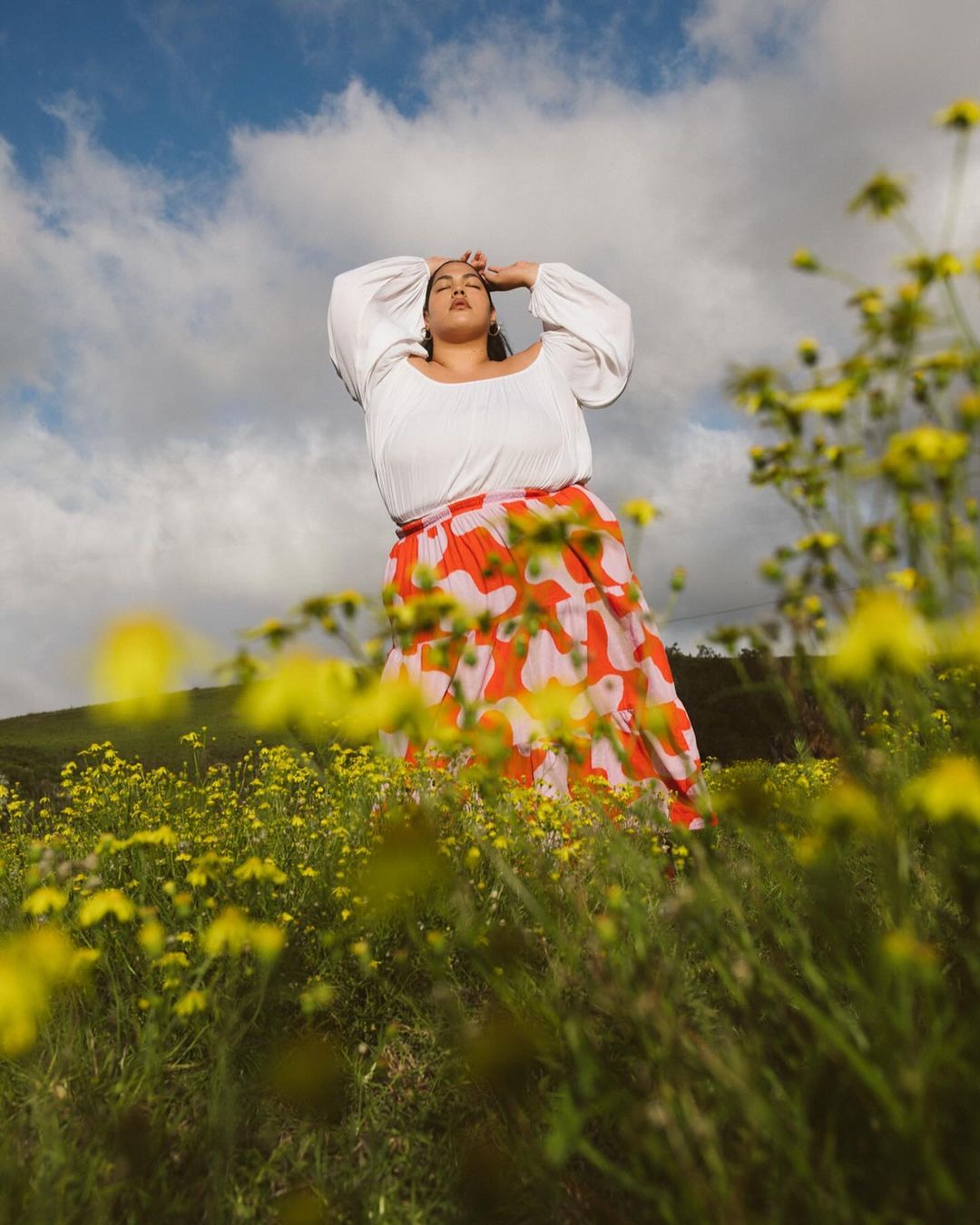 Woman in a field of yellow flowers wearing a white peasant blouse and a red floral skirt under a blue sky with clouds.