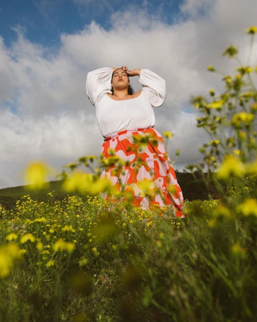 Woman in a field of yellow flowers wearing a white peasant blouse and a red floral skirt under a blue sky with clouds.