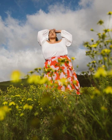 Woman in a field of yellow flowers wearing a white peasant blouse and a red floral skirt under a blue sky with clouds.
