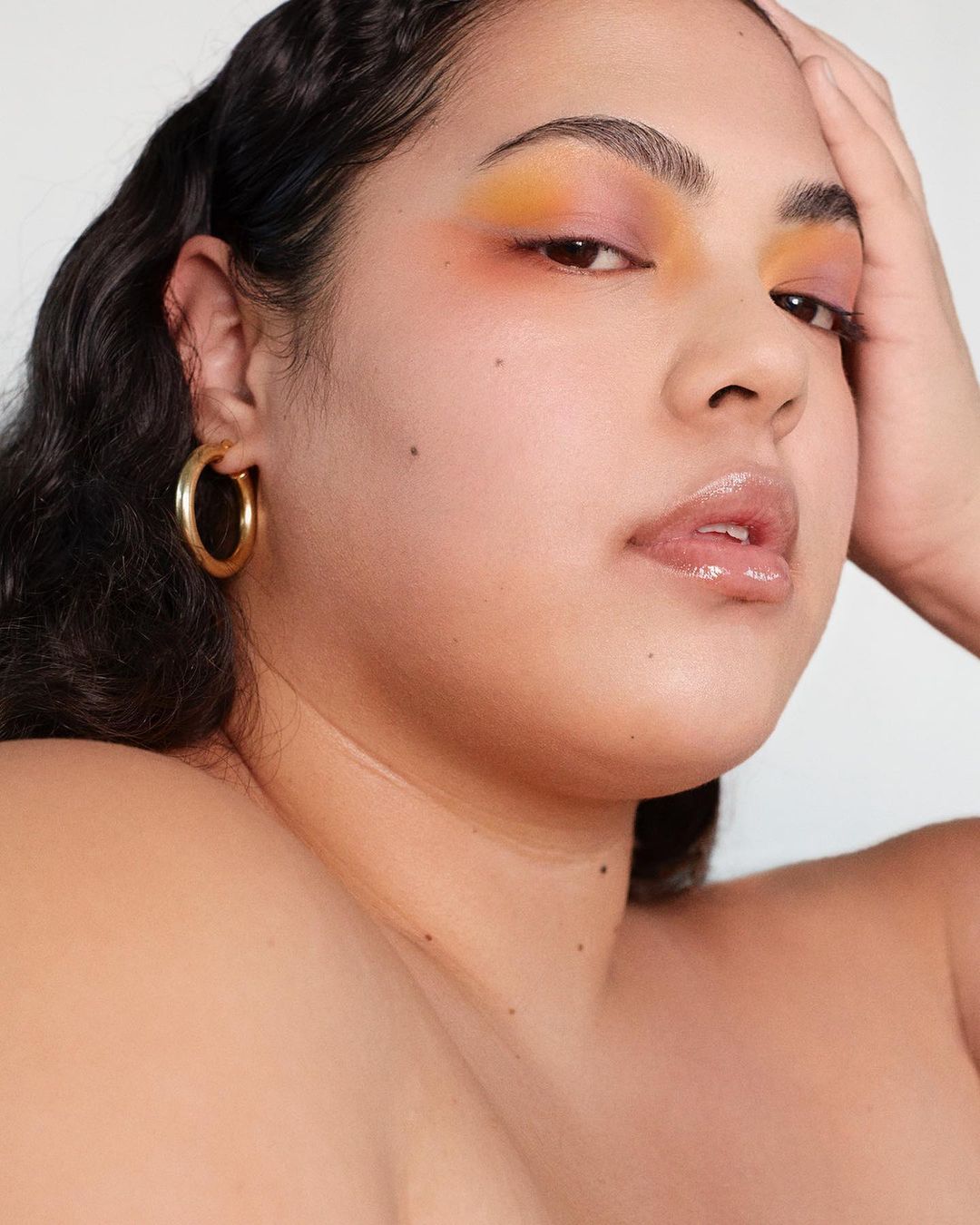Close-up portrait of a woman with gold hoop earrings and orange-toned eyeshadow posing indoors against a plain white background.