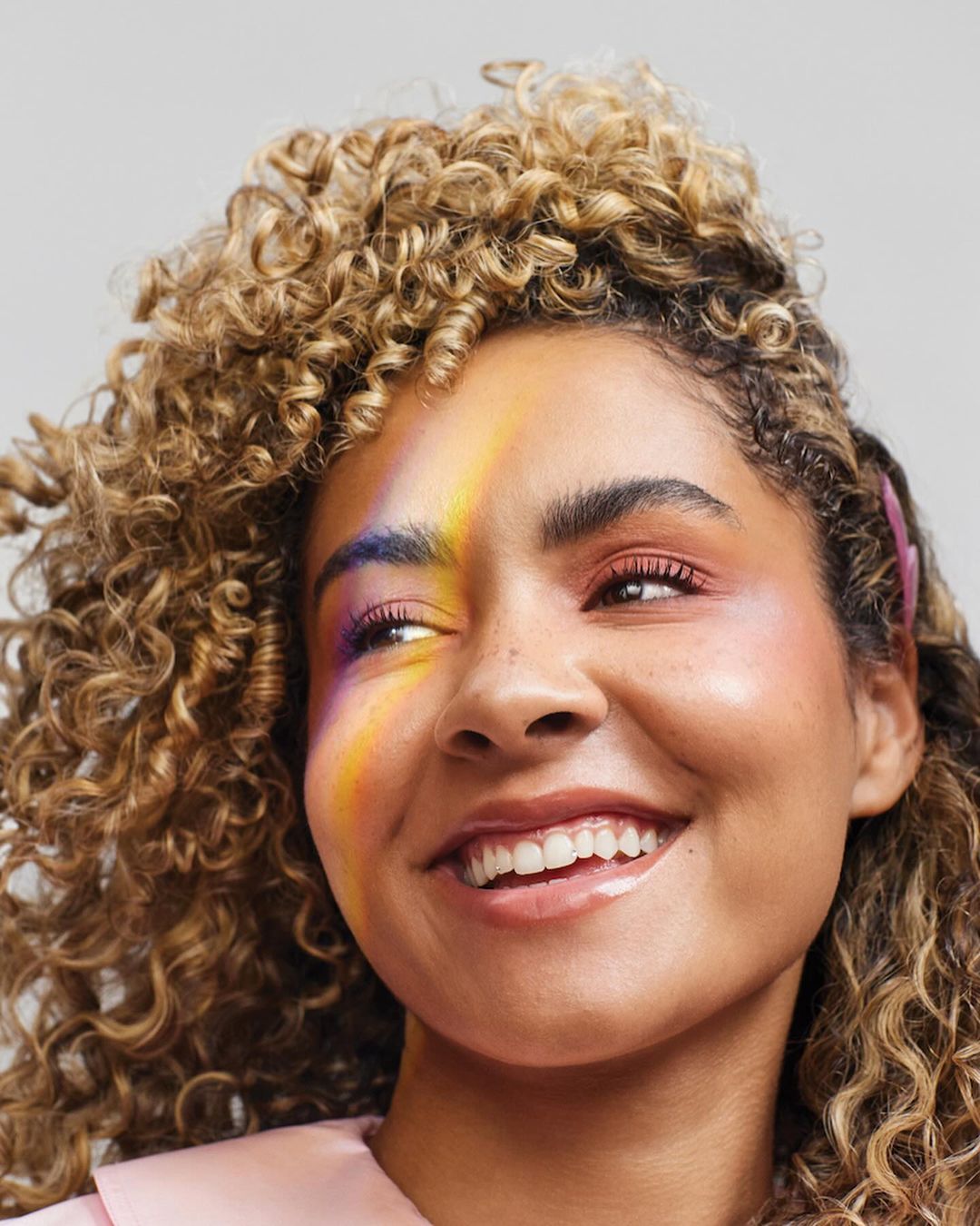 Close-up of an adult female smiling with curly blonde hair and rainbow makeup and wearing a pink shirt against a grey background.