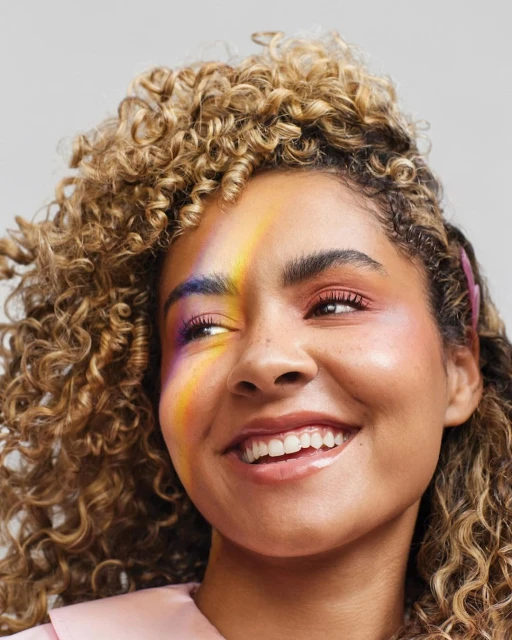Close-up of an adult female smiling with curly blonde hair and rainbow makeup and wearing a pink shirt against a grey background.