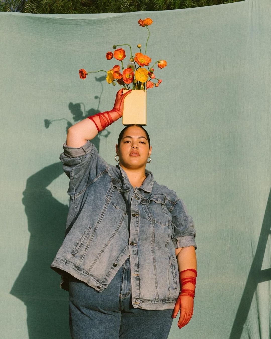 Woman in denim jacket balances a vase of orange poppies on her head, wearing sheer red gloves, standing against a solid teal background.