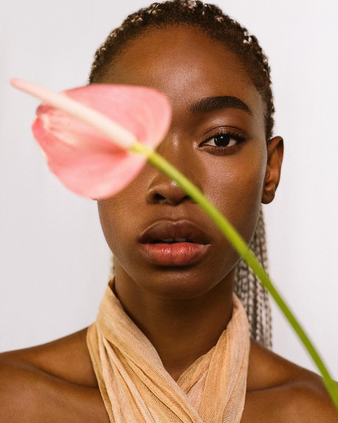 Close-up portrait of a young Black woman with braided hair, fair complexion, and light scarf, posing with a pink Anthurium flower.