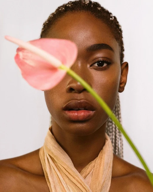 Close-up portrait of a young Black woman with braided hair, fair complexion, and light scarf, posing with a pink Anthurium flower.