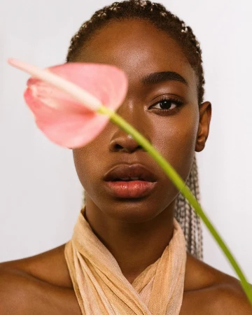 Close-up portrait of a young Black woman with braided hair, fair complexion, and light scarf, posing with a pink Anthurium flower.