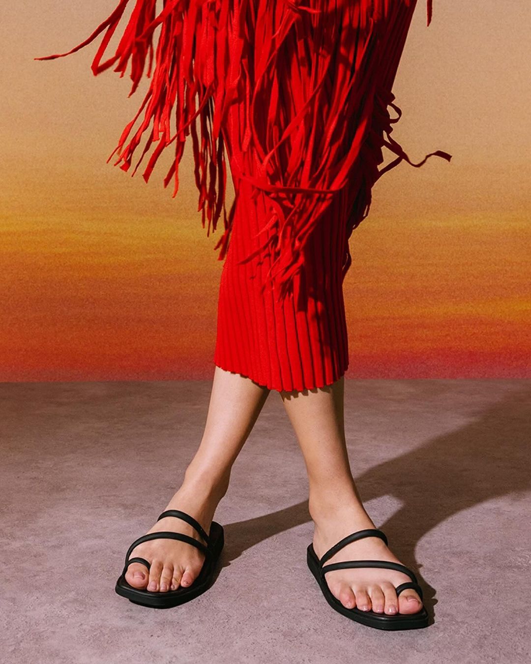 An adult female model is wearing black toe-ring sandals and a red fringe dress, posing against a warm gradient background in a studio.
