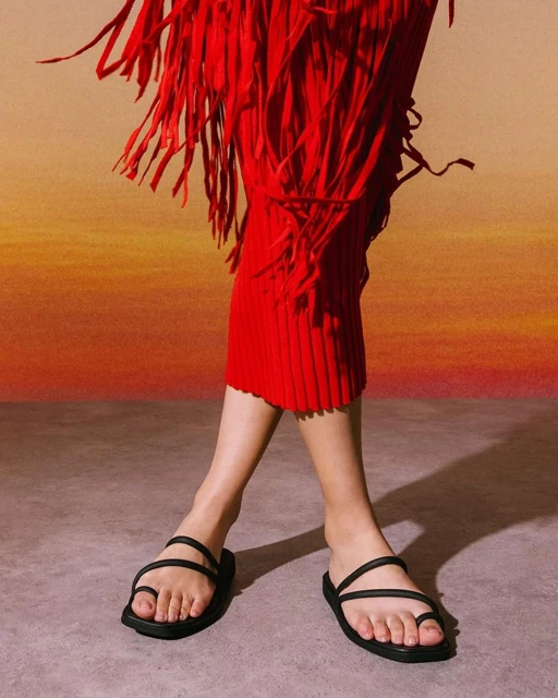 An adult female model is wearing black toe-ring sandals and a red fringe dress, posing against a warm gradient background in a studio.