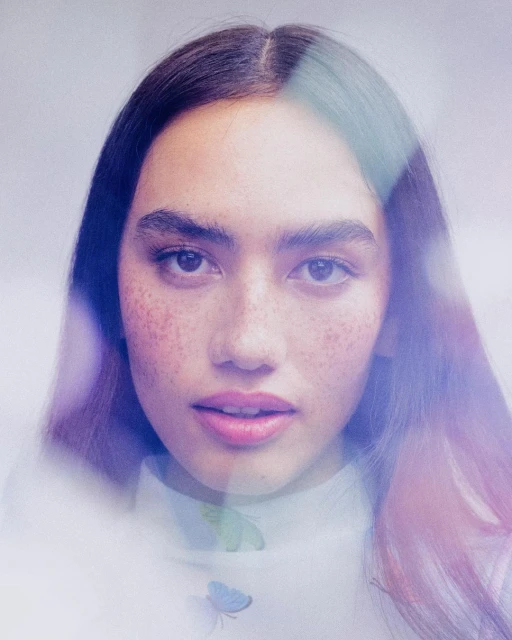 Close-up portrait of a young adult female with freckles, looking at the camera with a neutral facial expression. She is wearing a white top.