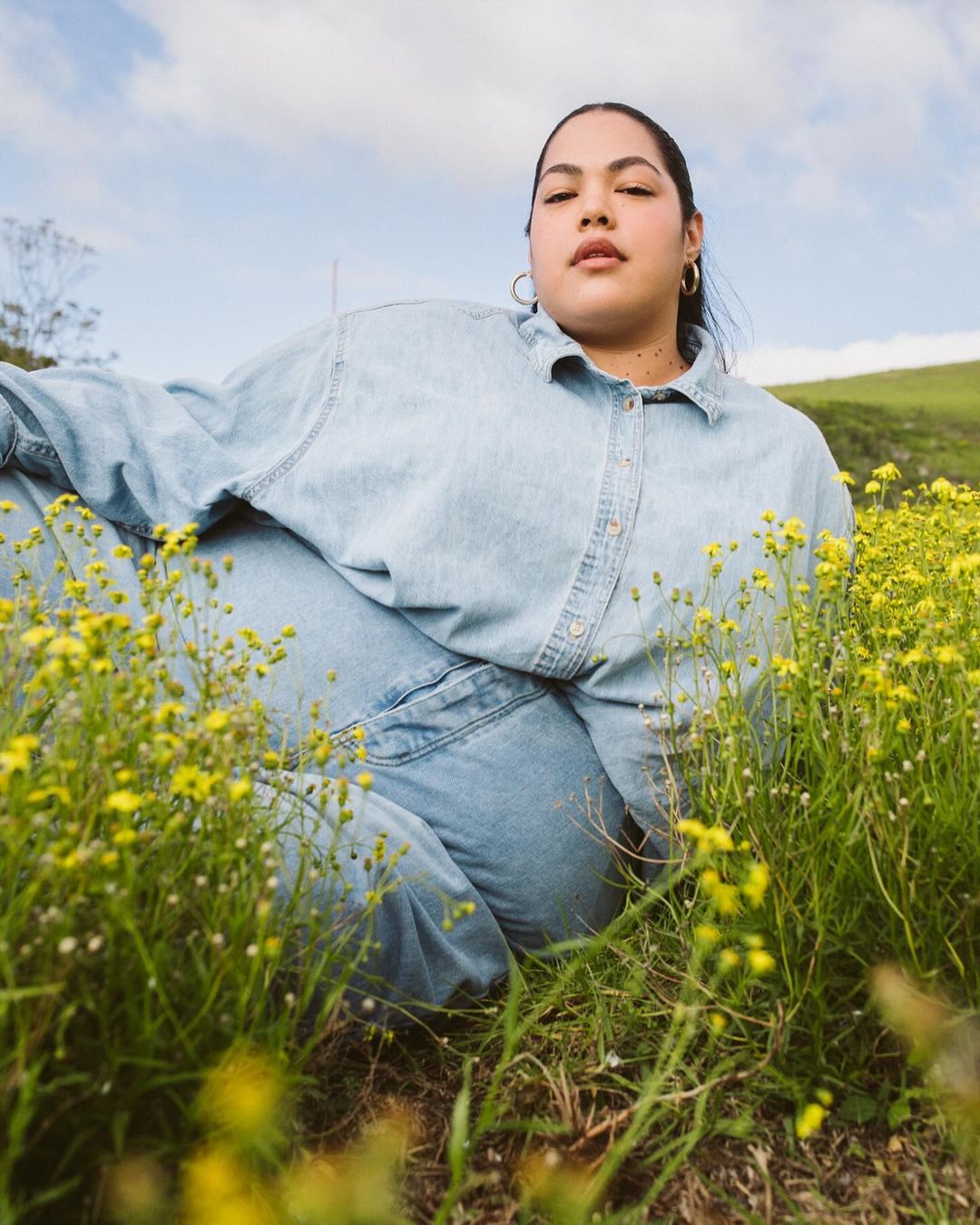 Woman lies in a field of yellow flowers, wearing denim clothing and gold hoop earrings, relaxed pose, with a green hill in the background.