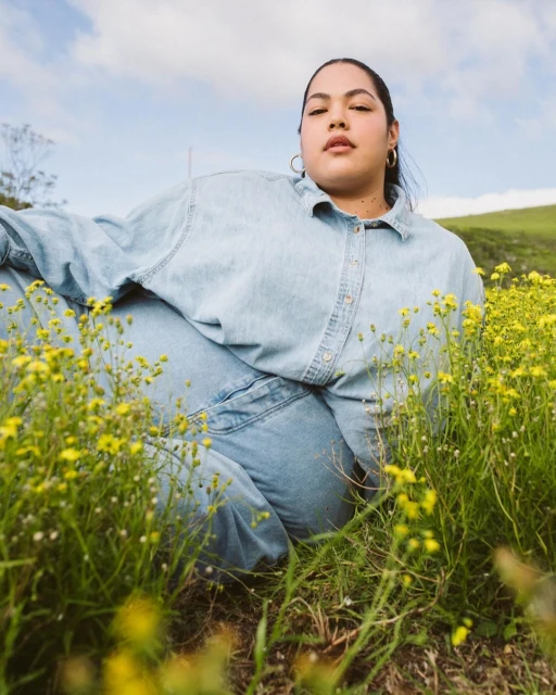 Woman lies in a field of yellow flowers, wearing denim clothing and gold hoop earrings, relaxed pose, with a green hill in the background.