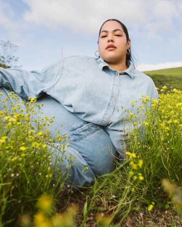 Woman lies in a field of yellow flowers, wearing denim clothing and gold hoop earrings, relaxed pose, with a green hill in the background.