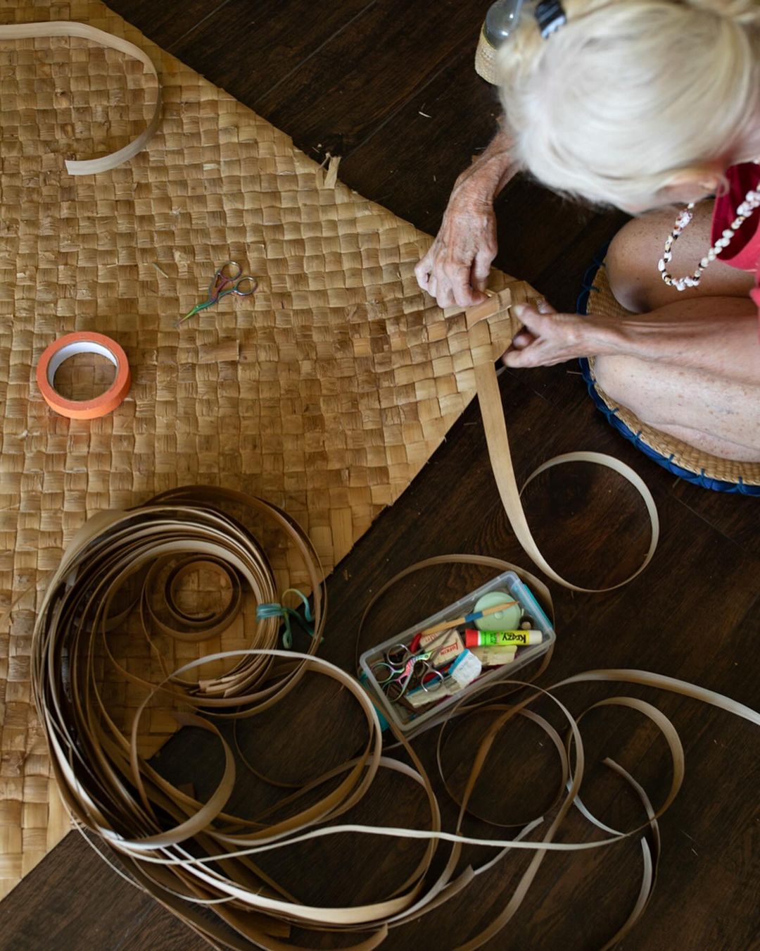 Overhead shot of an elderly woman weaving a mat with strips of tan material, surrounded by tools and supplies on a wood surface.