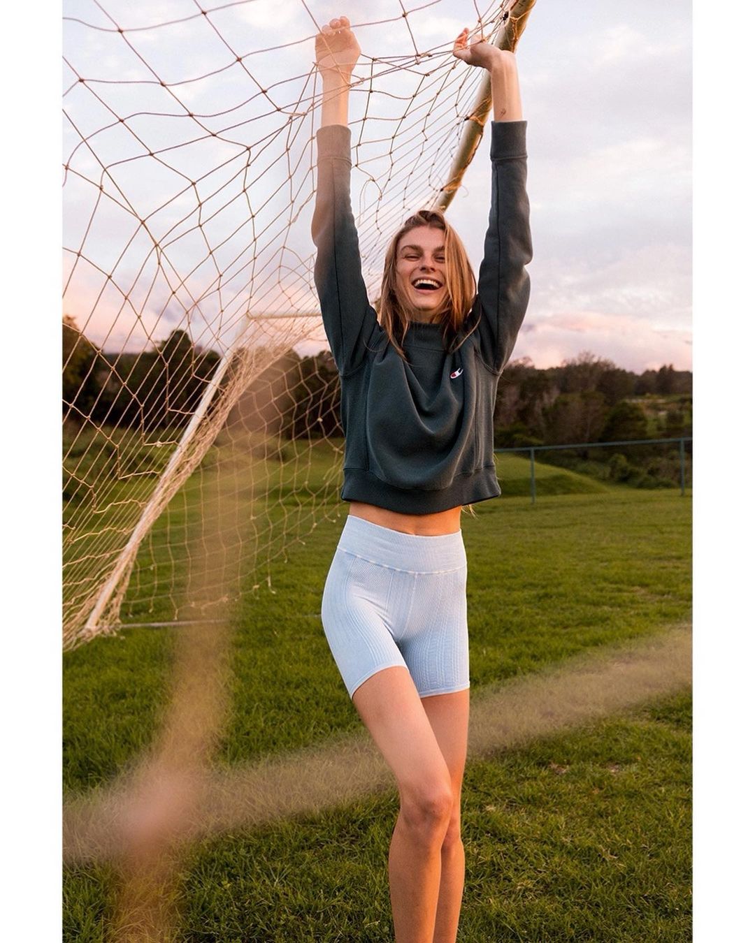 Young woman in athletic wear smiles while hanging from a soccer net in an outdoor field at dusk, creating a playful, active image.
