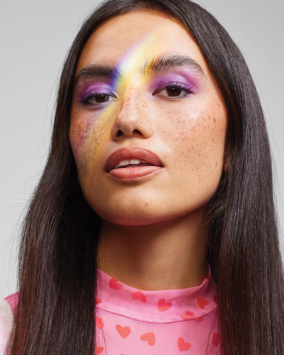 Portrait of a woman with freckles wearing purple eyeshadow and a rainbow light across her face against a grey backdrop.