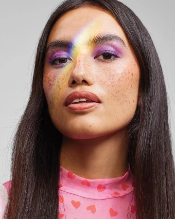 Portrait of a woman with freckles wearing purple eyeshadow and a rainbow light across her face against a grey backdrop.