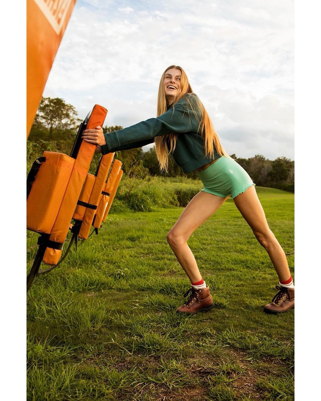 Athletic woman presses against orange padded equipment on a green field with a blue sky in an active outdoor setting.