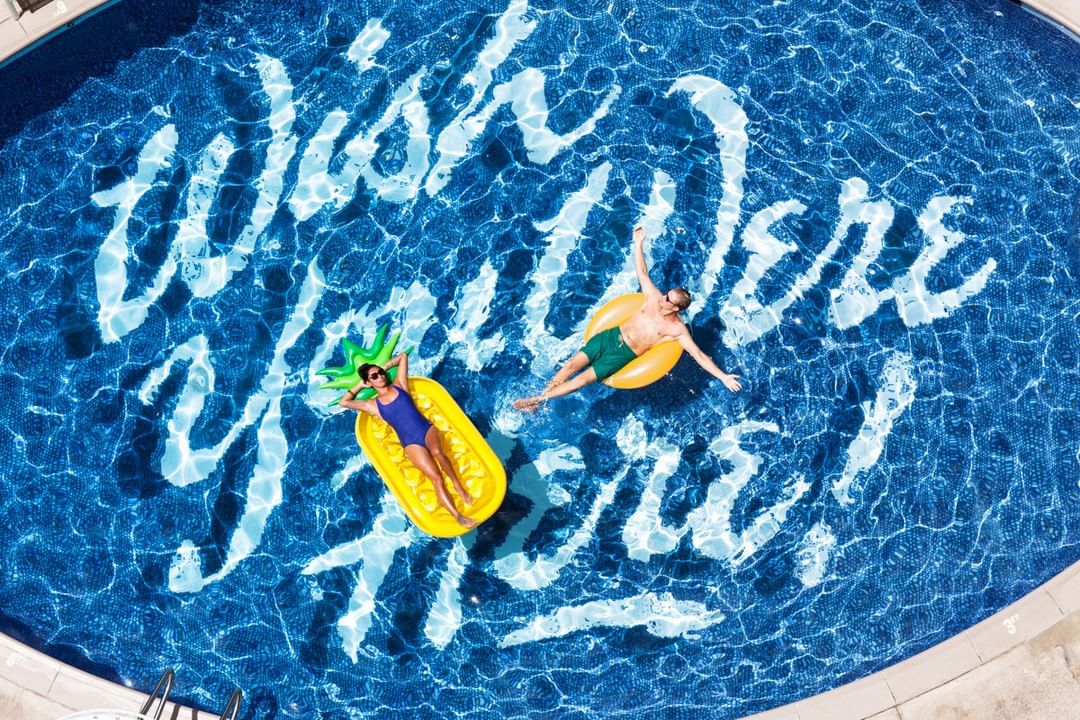 Aerial view of a woman on a pineapple float and a man on a donut float in a swimming pool with the text 'Wish You Were Here'.