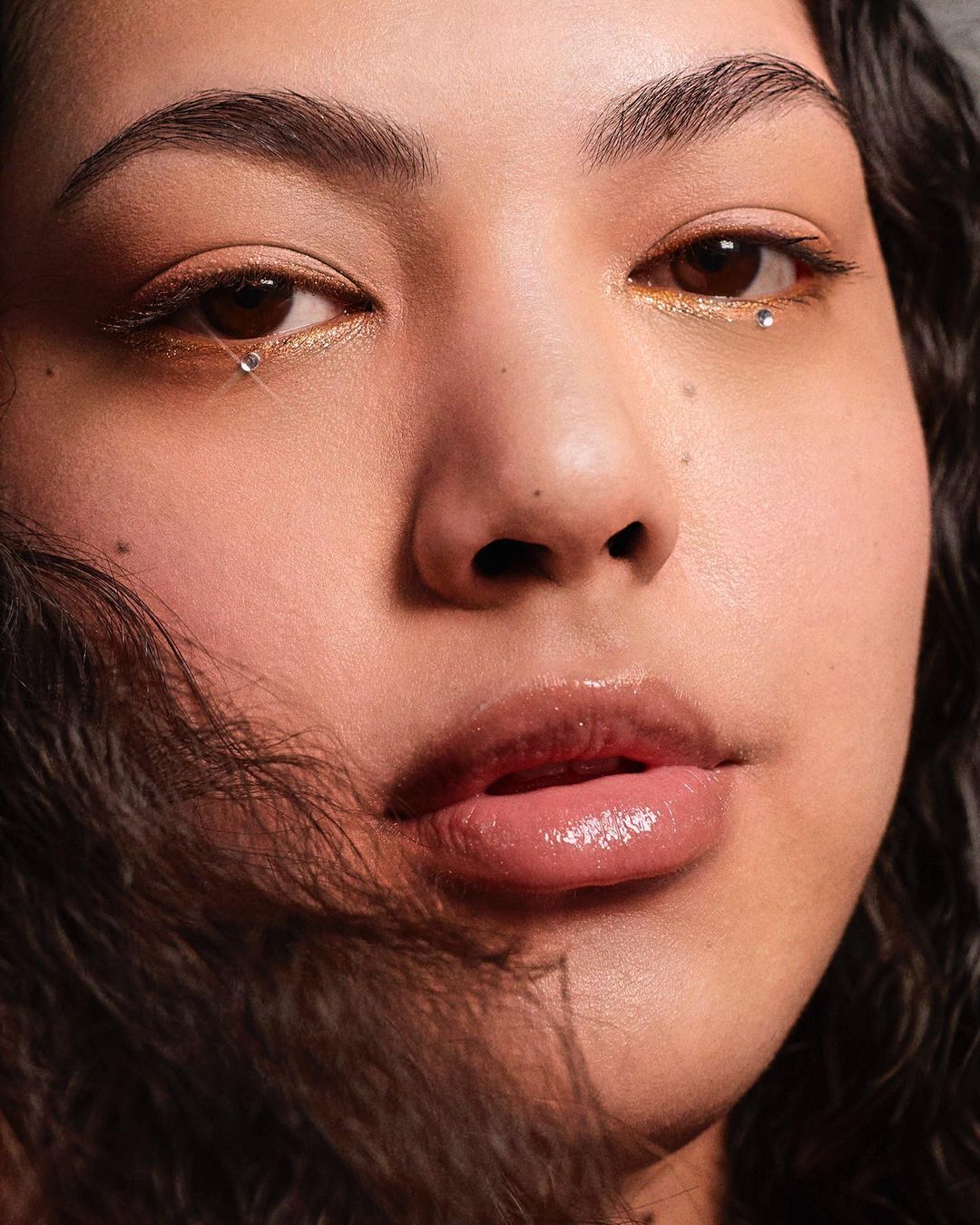 Close-up portrait of a young woman with golden eye makeup and rhinestones under the eyes, soft skin, and curly dark hair.