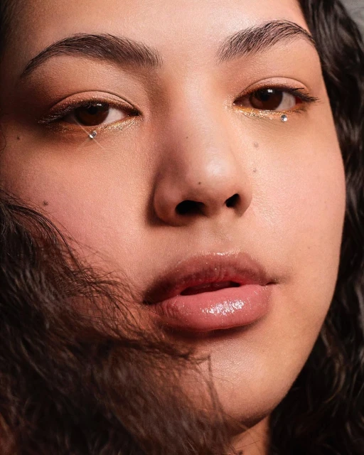 Close-up portrait of a young woman with golden eye makeup and rhinestones under the eyes, soft skin, and curly dark hair.