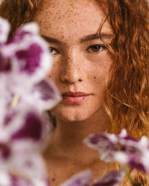 Portrait of a freckled woman with curly red hair, posing with purple orchids in an eye-level close-up shot, showcasing beauty and nature.