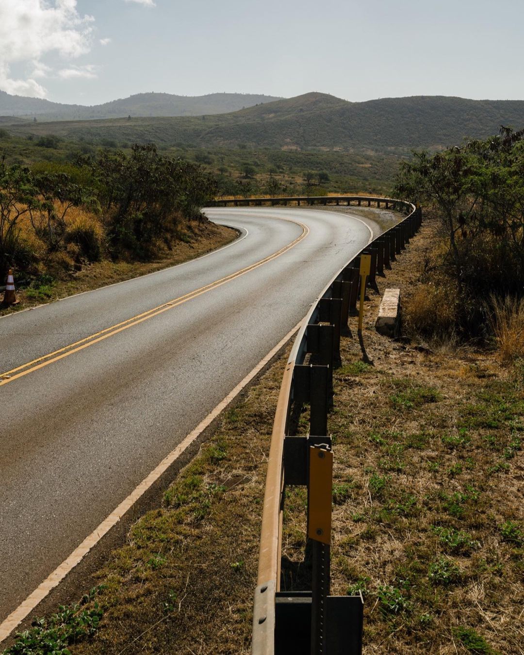 Winding road with a guardrail curves through a rural landscape under a bright sky, leading the eye through hills and greenery.