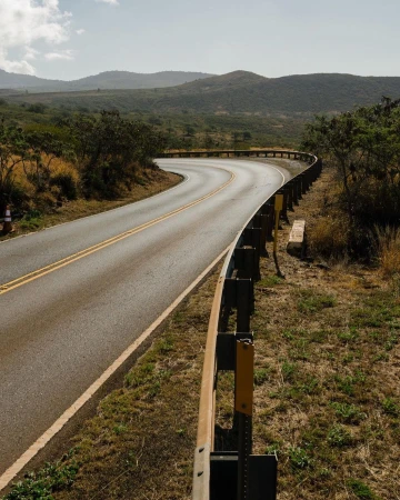 Winding road with a guardrail curves through a rural landscape under a bright sky, leading the eye through hills and greenery.