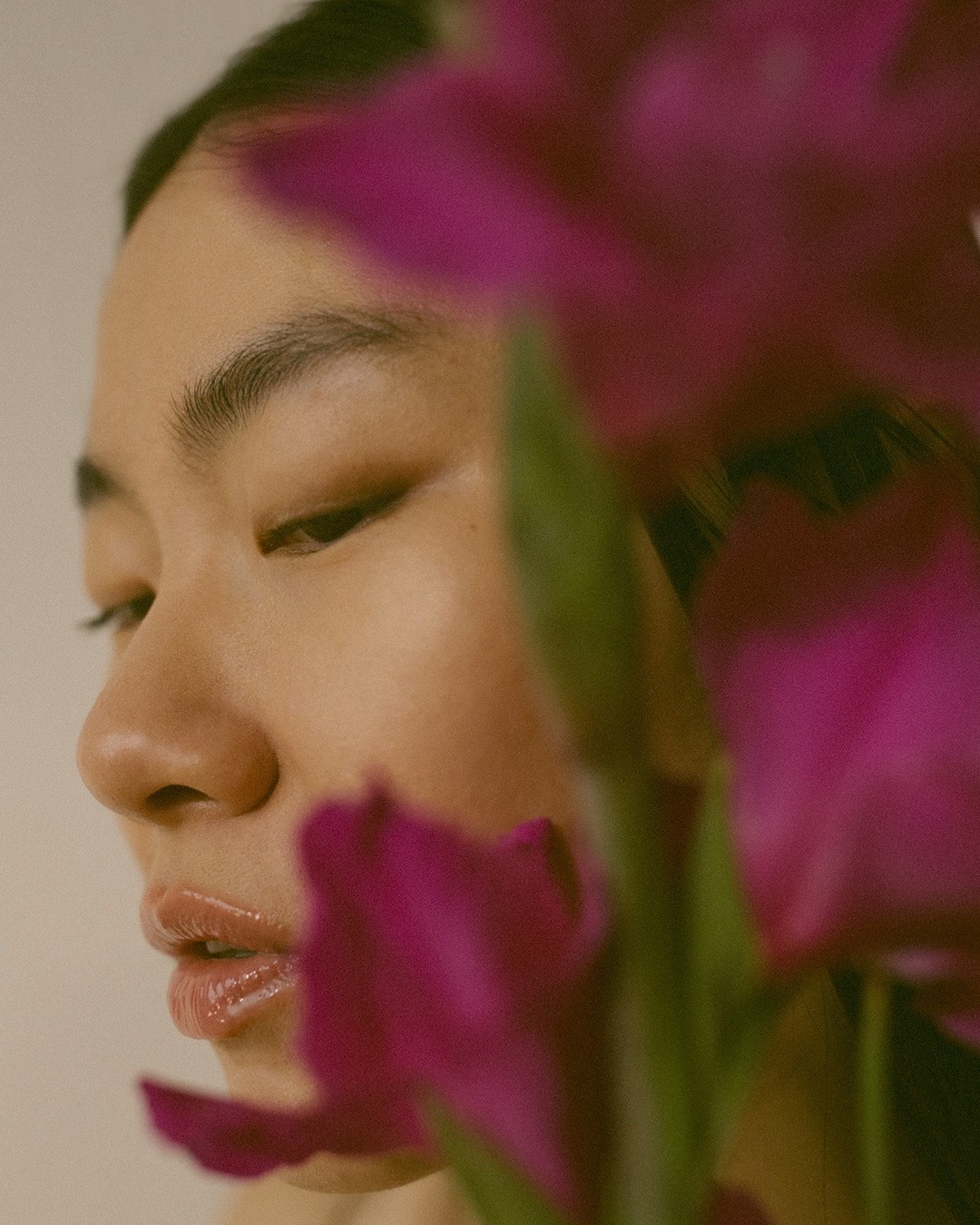 Close-up studio portrait of a young adult Asian woman with fair skin partially obscured by large purple flowers with green stems.