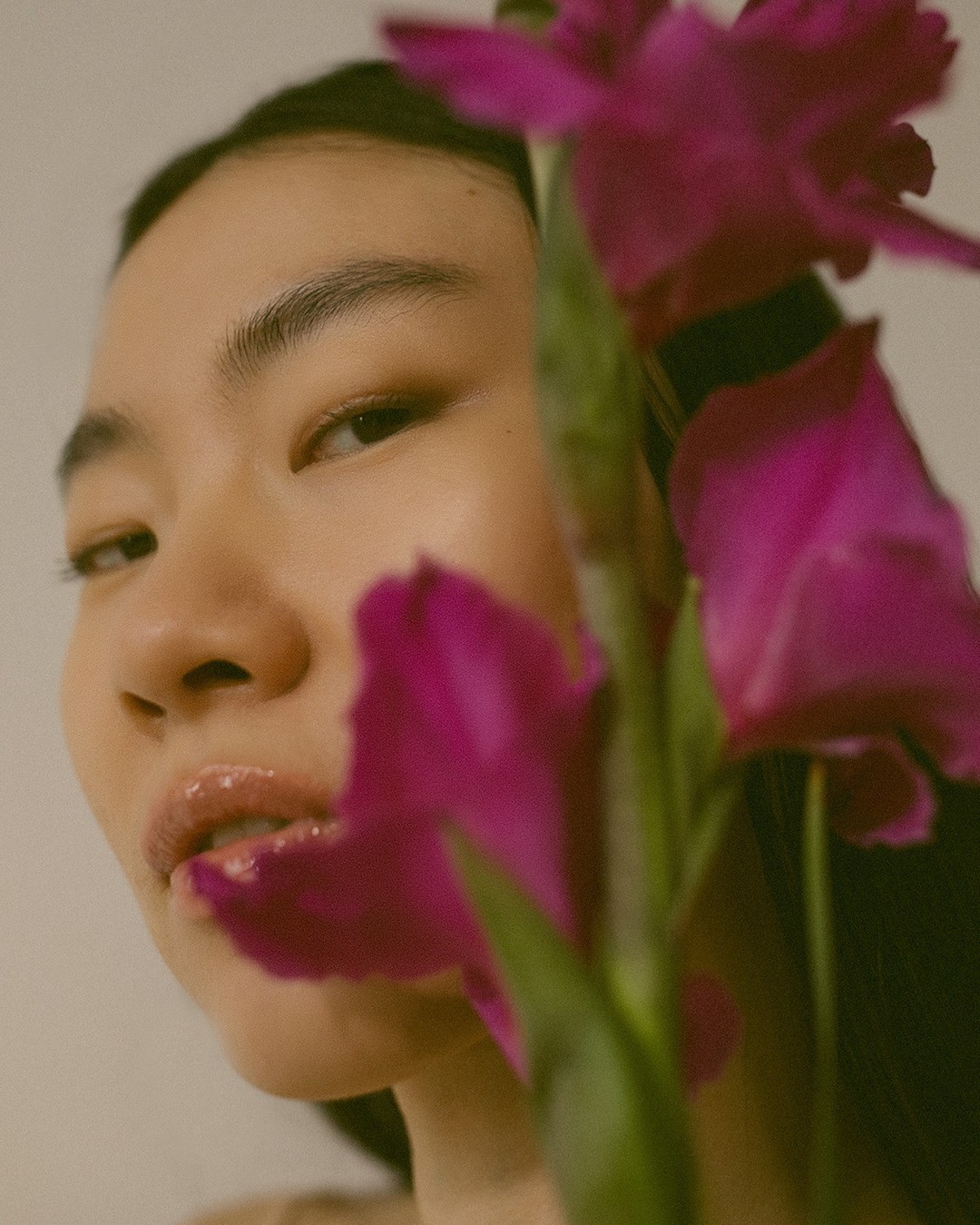 Close-up portrait of a young Asian woman with glossy lips partially obscured by magenta-colored gladiolus flowers against a light backdrop.